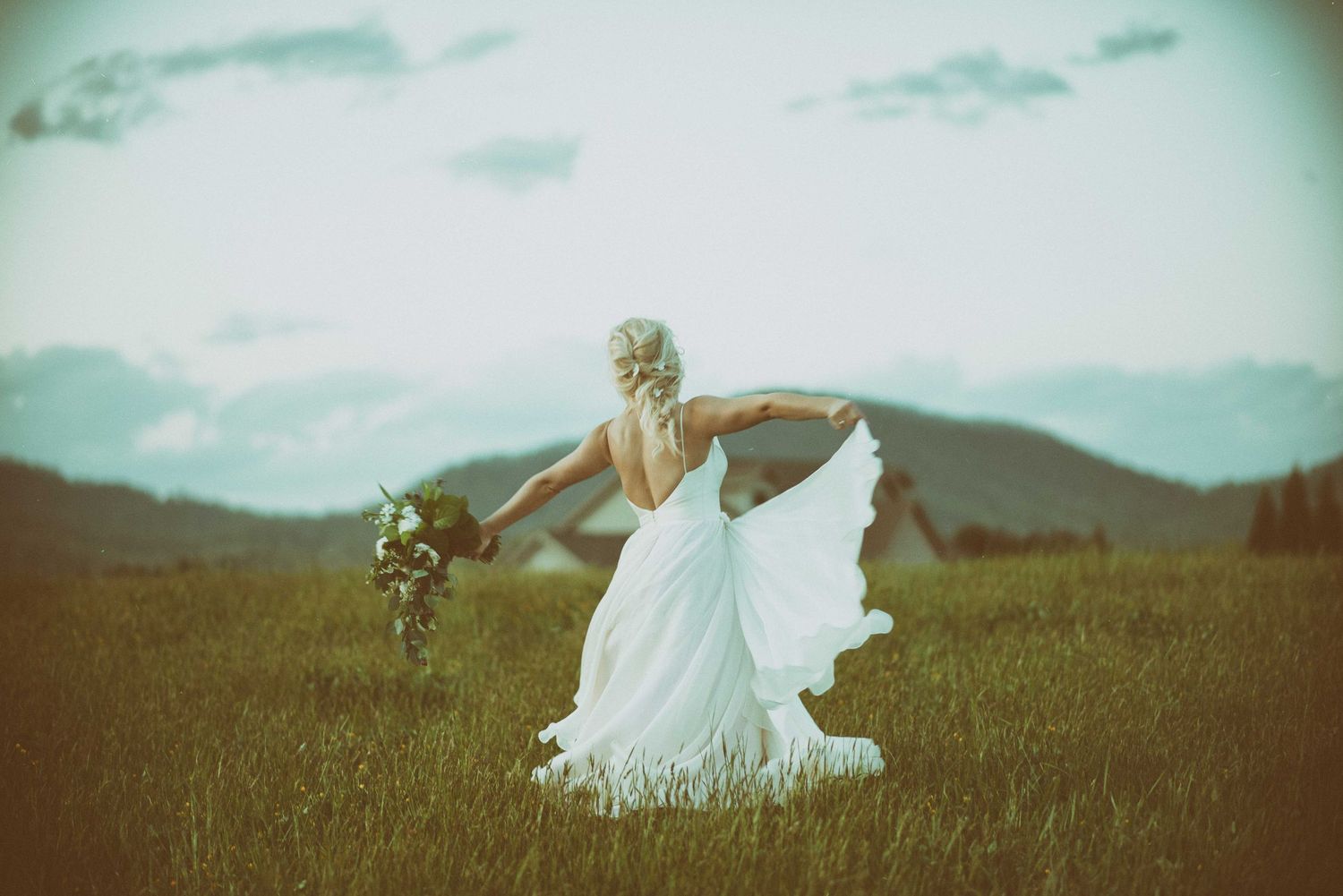 Bride in flowing white gown holding bouquet walks through mountain meadow on her wedding day.