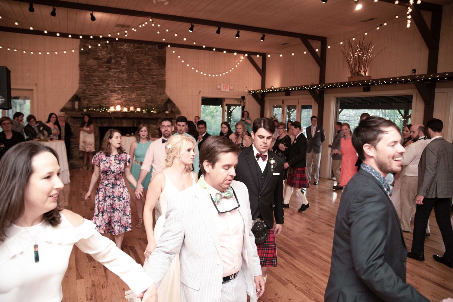 Wedding reception guests dancing in a rustic barn venue with string lights and stone fireplace.