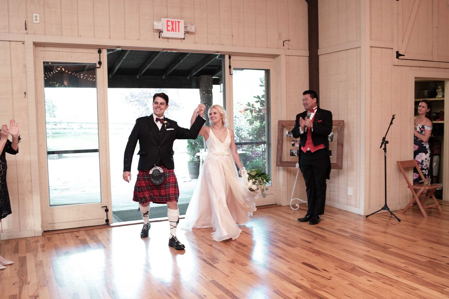 Groom in traditional Scottish kilt enters wedding reception with bride on hardwood dance floor.