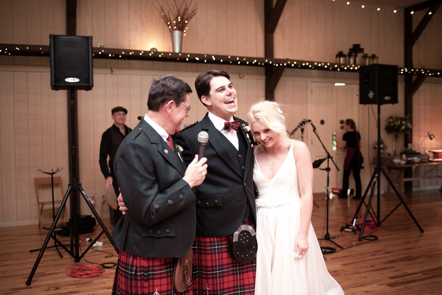 A groom in a kilt shares a microphone with the bride and groom at their wedding reception in a rustic barn venue.