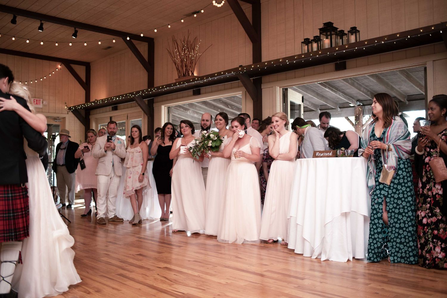 Bridesmaids in blush pink dresses line the dance floor at a rustic barn wedding reception with string lights.