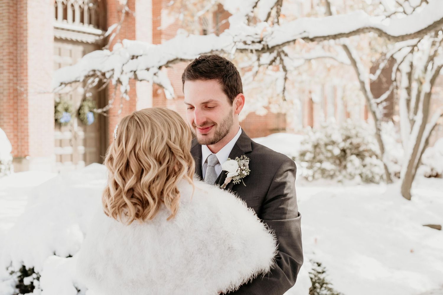Bride in white fur wrap and groom in gray suit share romantic moment in snowy winter wedding outdoor setting.