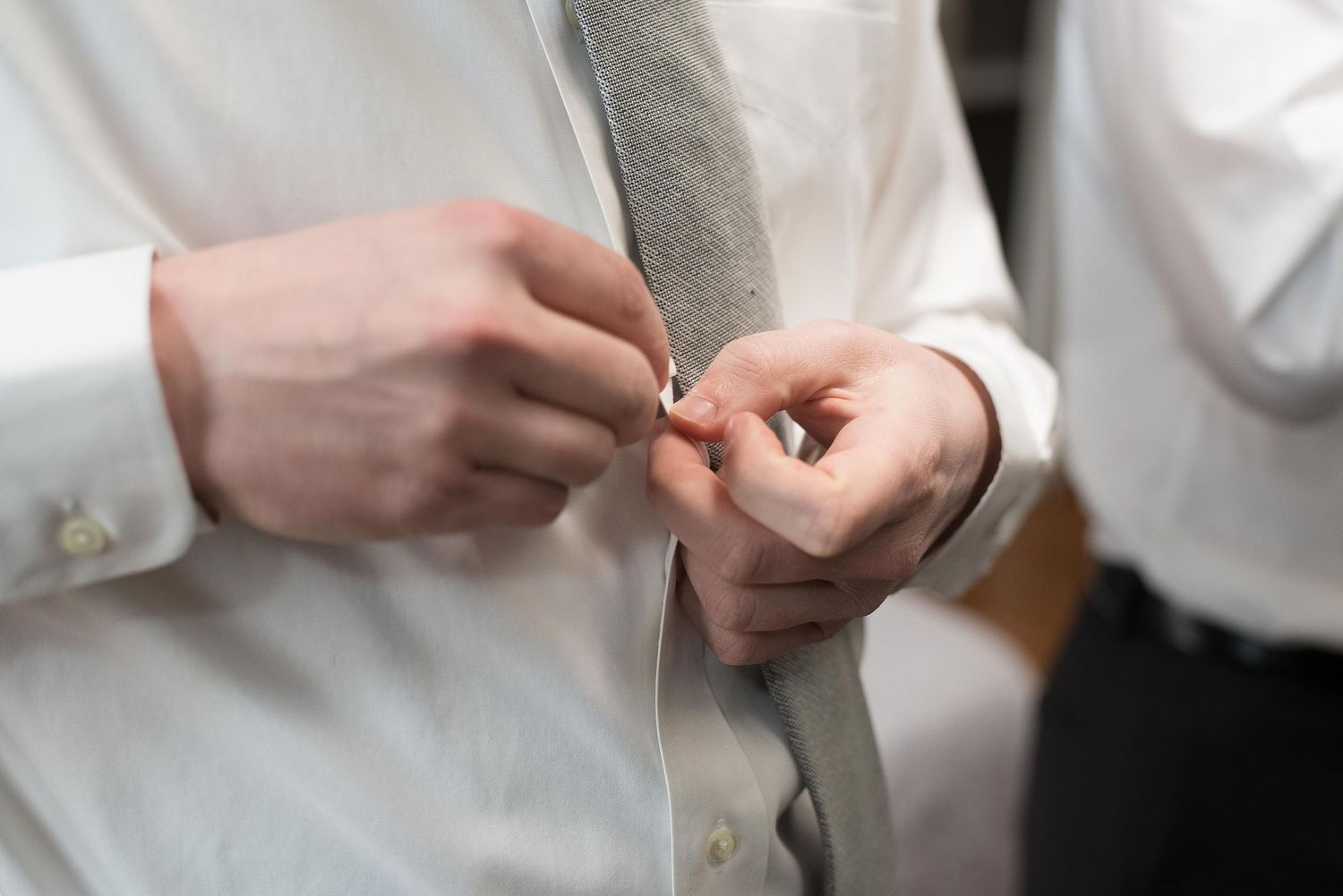 Man in white dress shirt fastening a gray tie, close-up of hands adjusting formal wedding attire.