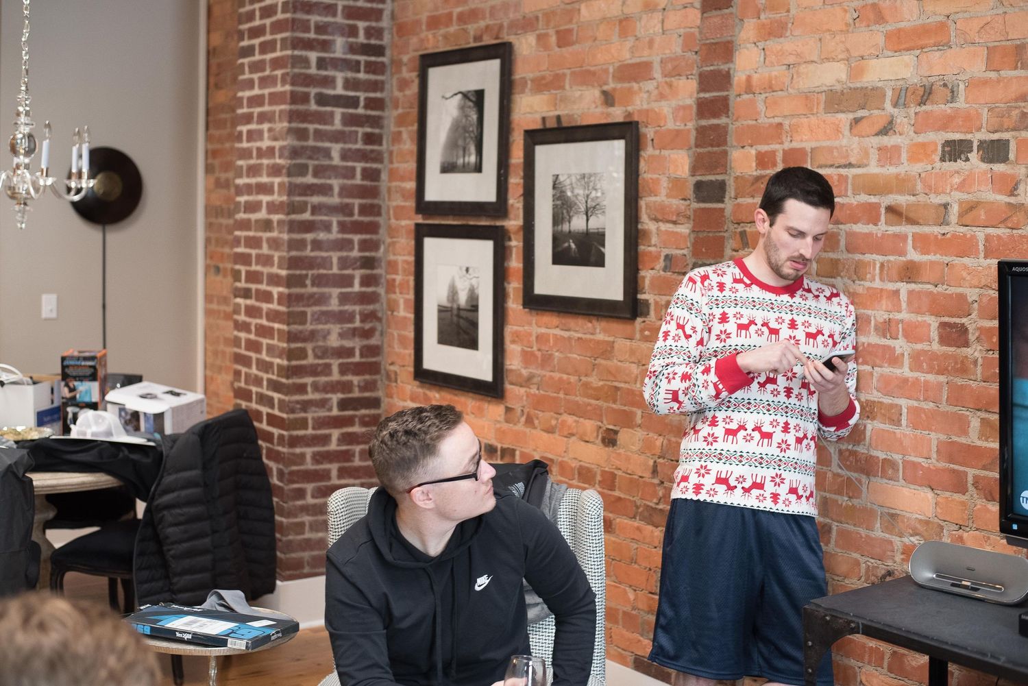 Two men in a brick-walled office, one wearing a festive Christmas sweater holding a phone.