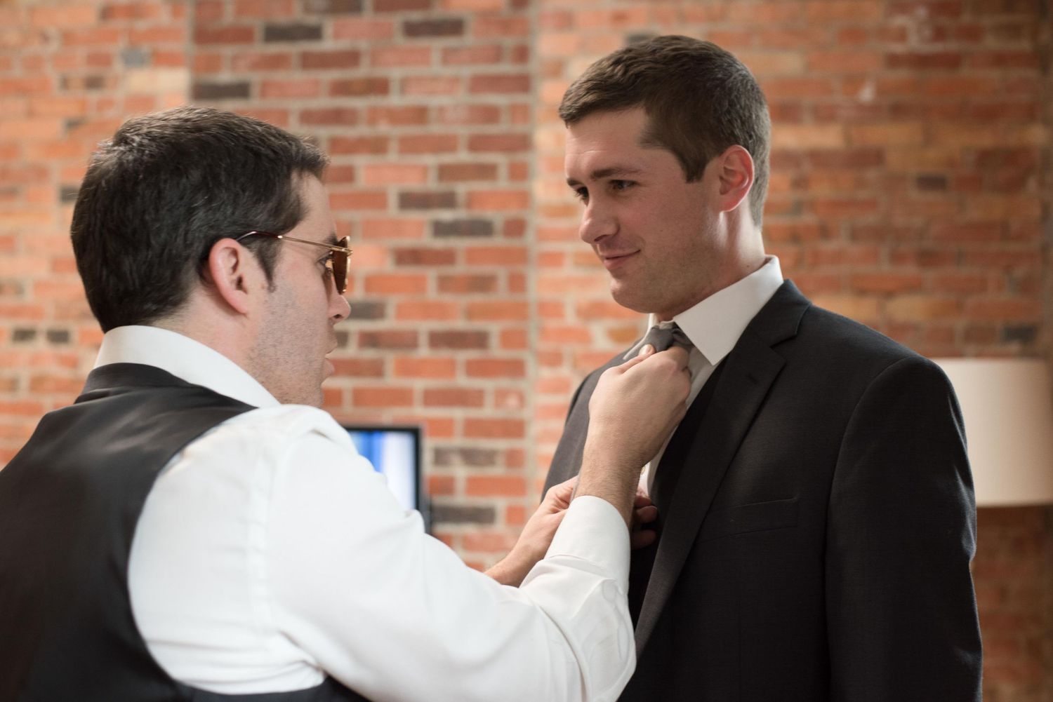 Man in vest adjusting tie of man in dark suit near brick wall.