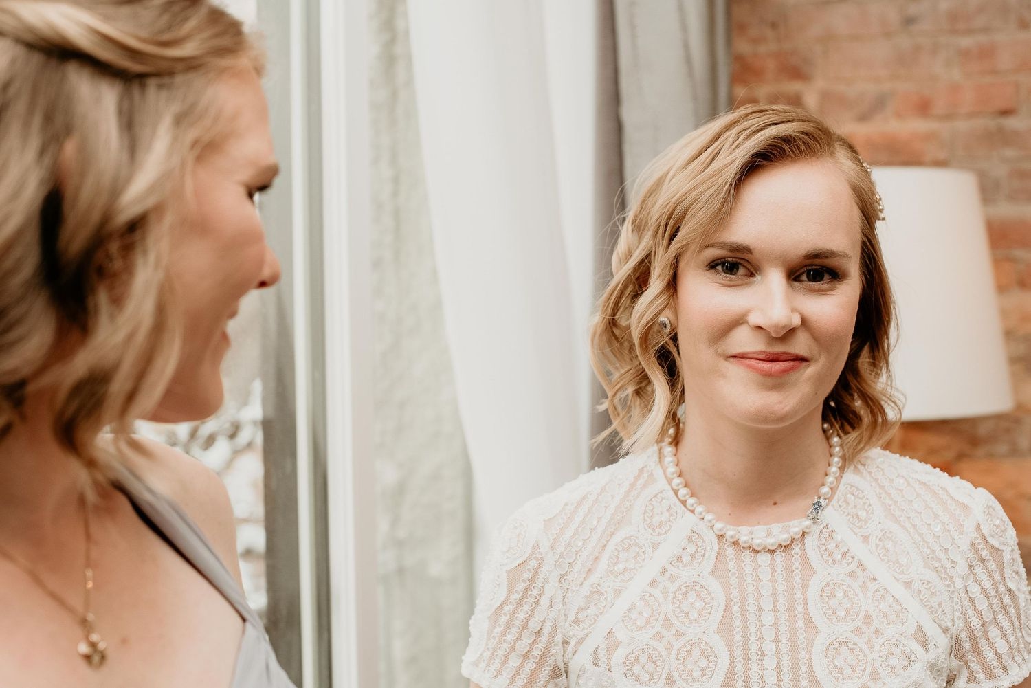 Bride and bridesmaid sharing a joyful moment, wearing elegant white lace dress and pearl necklace.