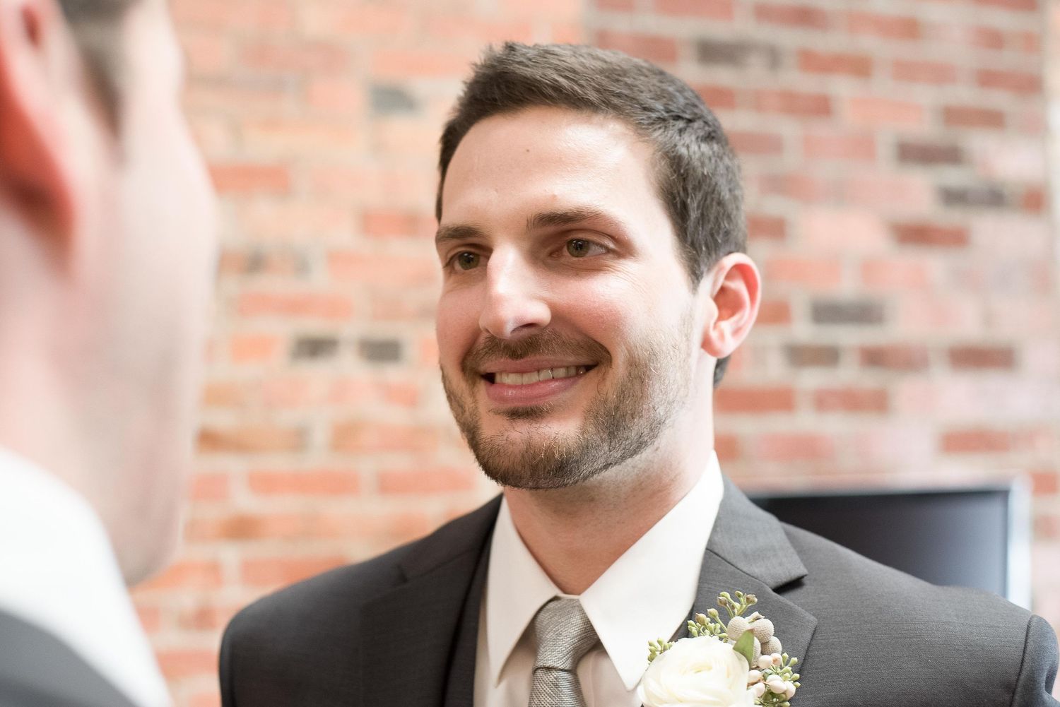 Smiling groom in gray suit with boutonniere at wedding ceremony near brick wall.