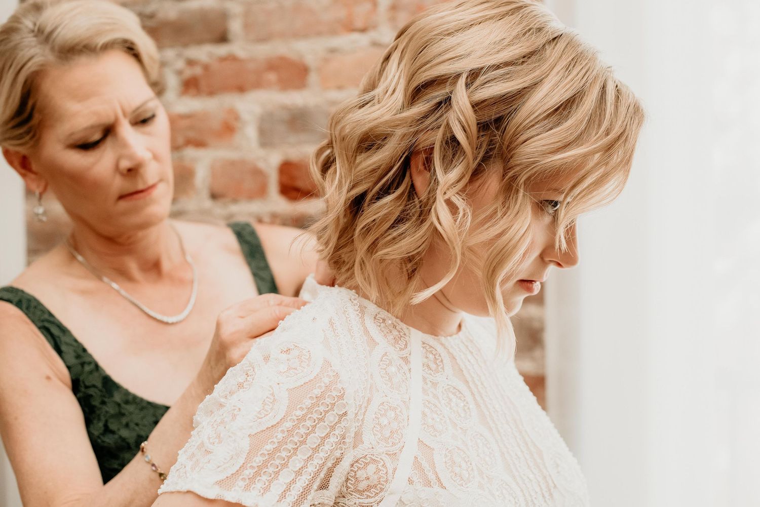 Woman in green dress carefully fastening the back of a bride's lace wedding dress indoors.