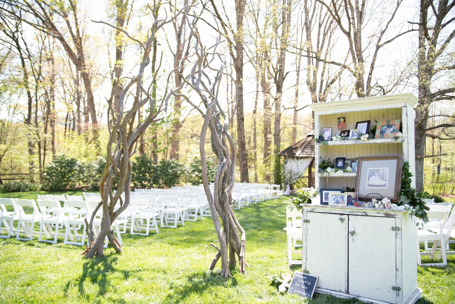 Outdoor wedding ceremony setup with white chairs on green lawn, bare trees, and a vintage display cabinet with framed photos.