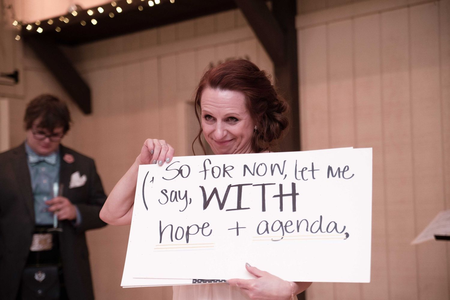 Woman holding a handwritten sign reading 'So for now, let me say, WITH hope + agenda' at a wedding reception.