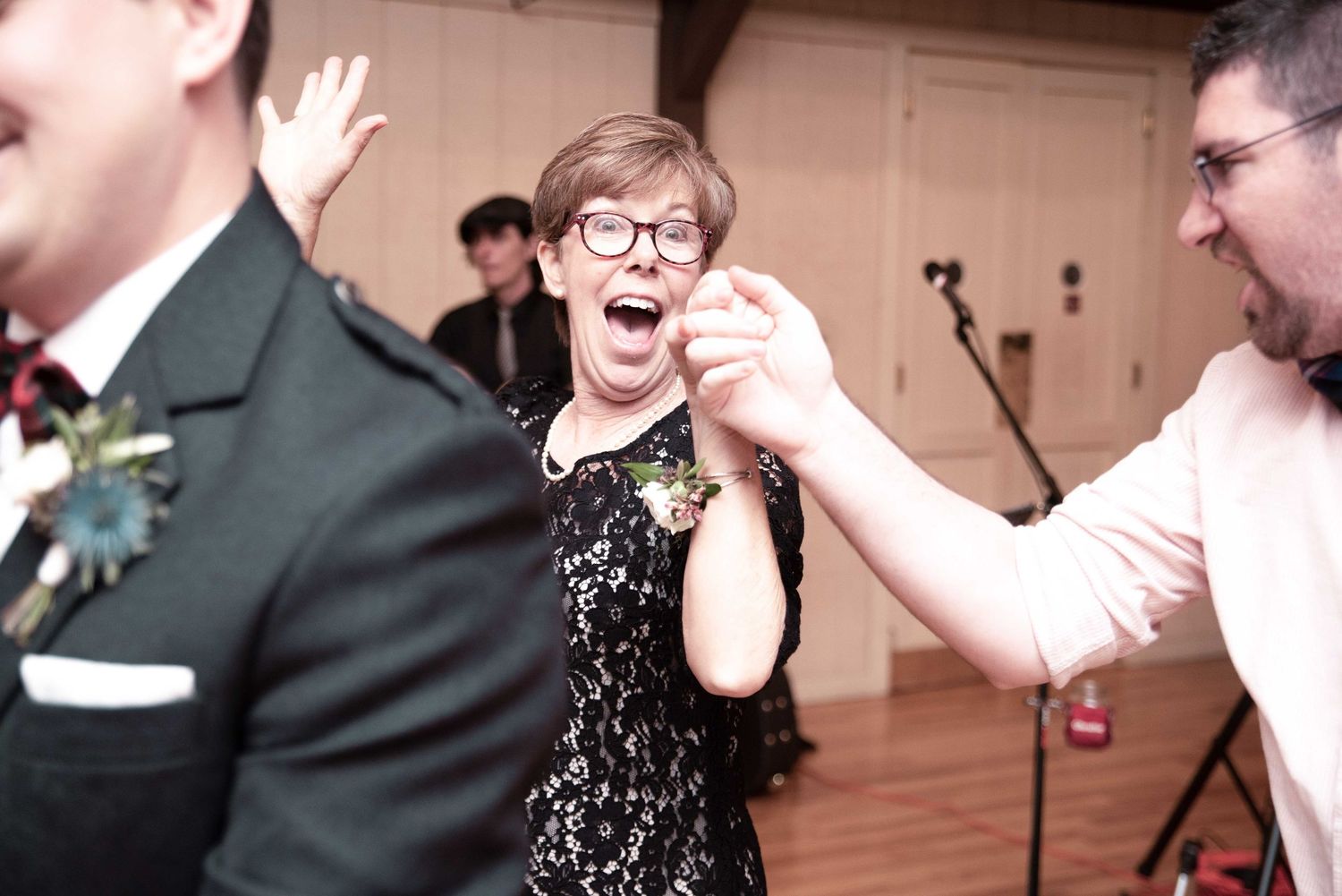 Woman in black lace dress dancing joyfully at a wedding reception, laughing and holding hands with guests.