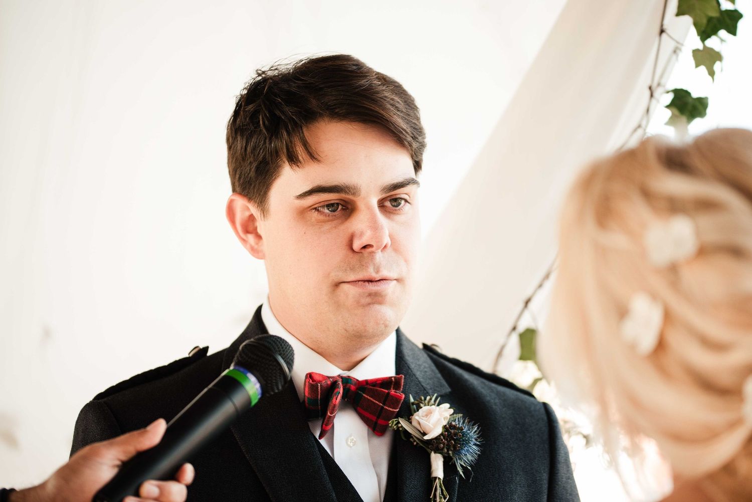 A groom in a dark suit and red tartan kilt listens intently during his wedding ceremony vows.