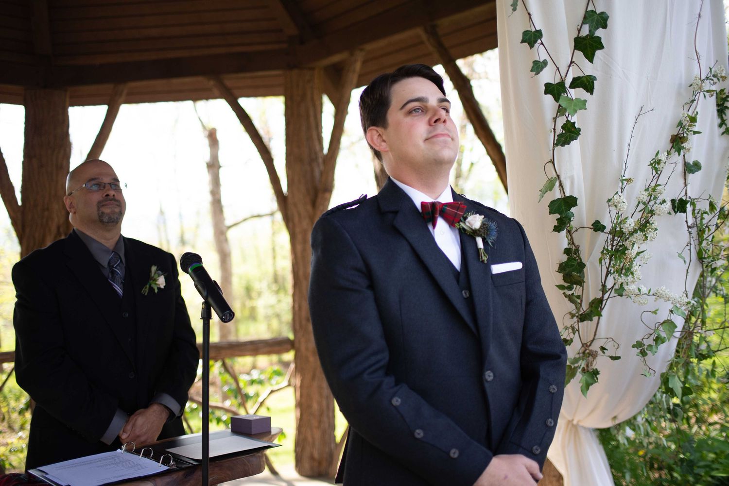 Groom in navy suit with burgundy bow tie stands at outdoor wedding altar, looking emotional as bride approaches.