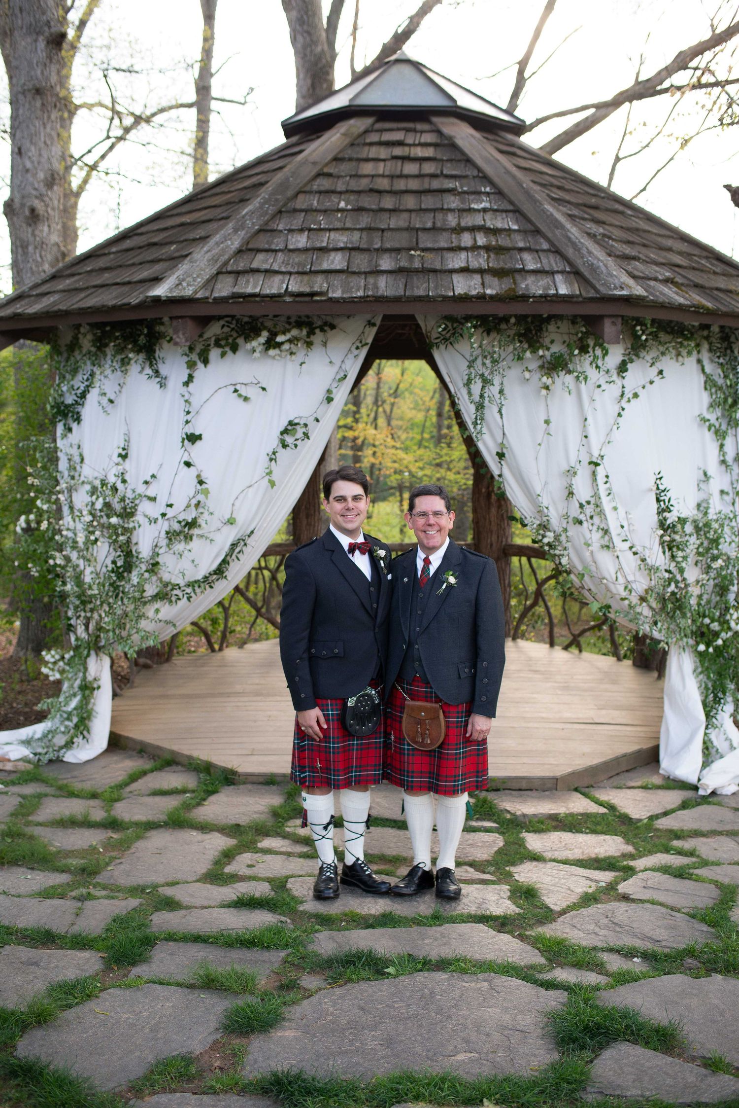 Two grooms in kilts and suits pose before a decorated gazebo with white drapes and greenery at an outdoor wedding.