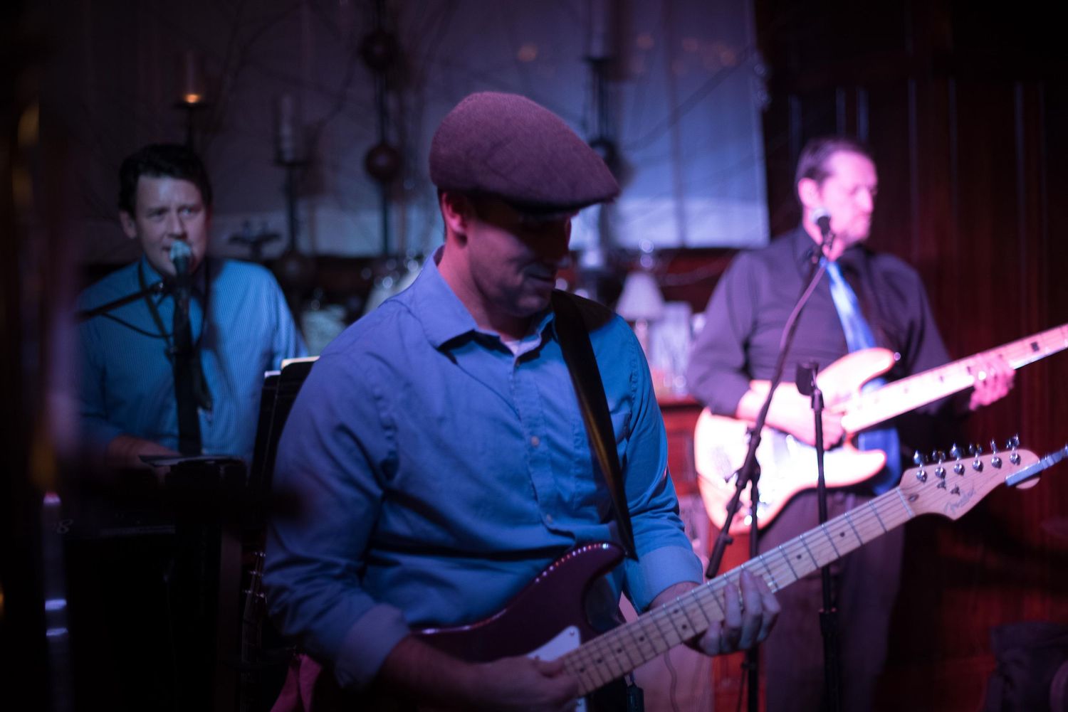 Three musicians performing live, with a guitarist in a flat cap playing electric guitar under warm stage lighting.