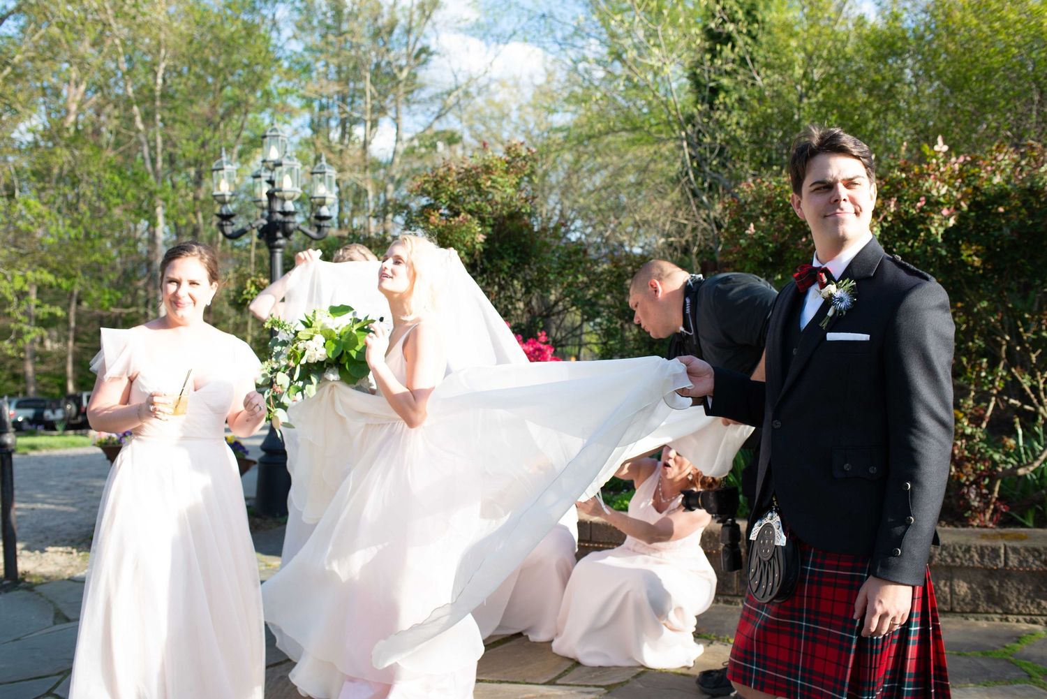 Bride in white gown with bridesmaids and groom in red tartan kilt at outdoor wedding ceremony.