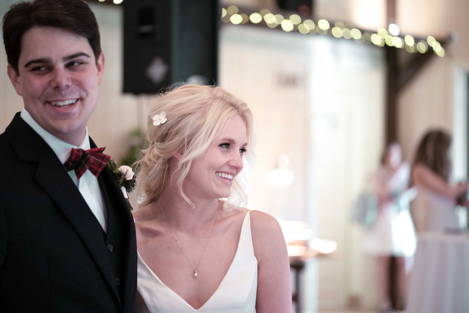 A smiling bride and groom in formal wedding attire pose together at their elegant reception venue with warm string lights.