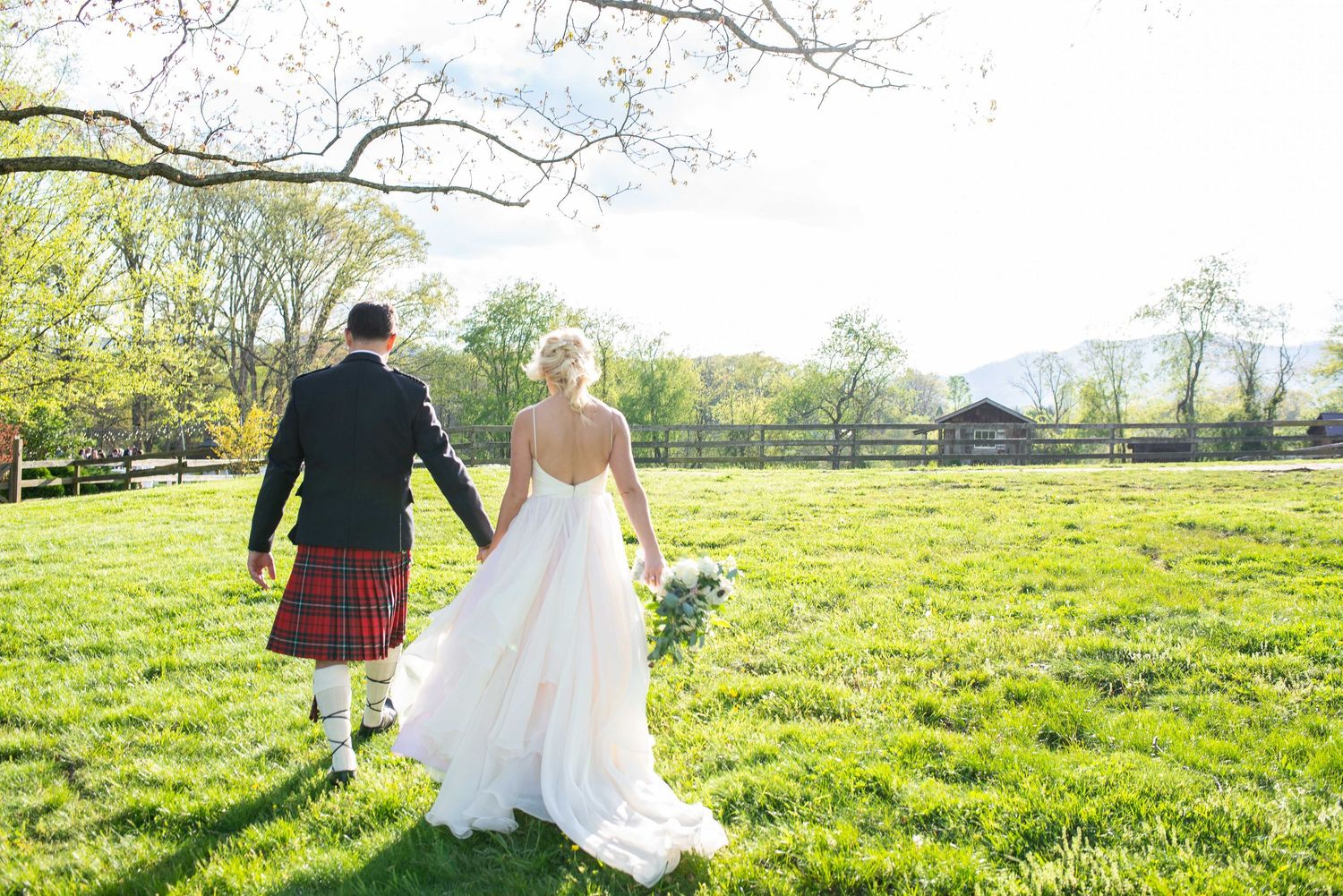 Bride in white gown and groom in red tartan kilt hold hands walking across green field at outdoor wedding.