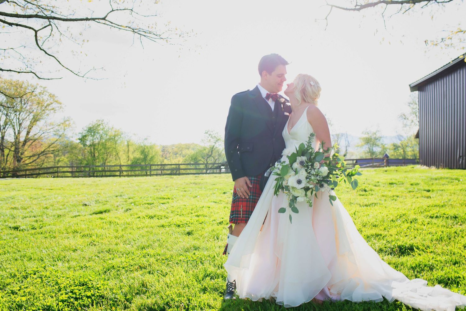 Bride and groom share a romantic moment on a sunlit green lawn, bride holding a lush white floral bouquet.