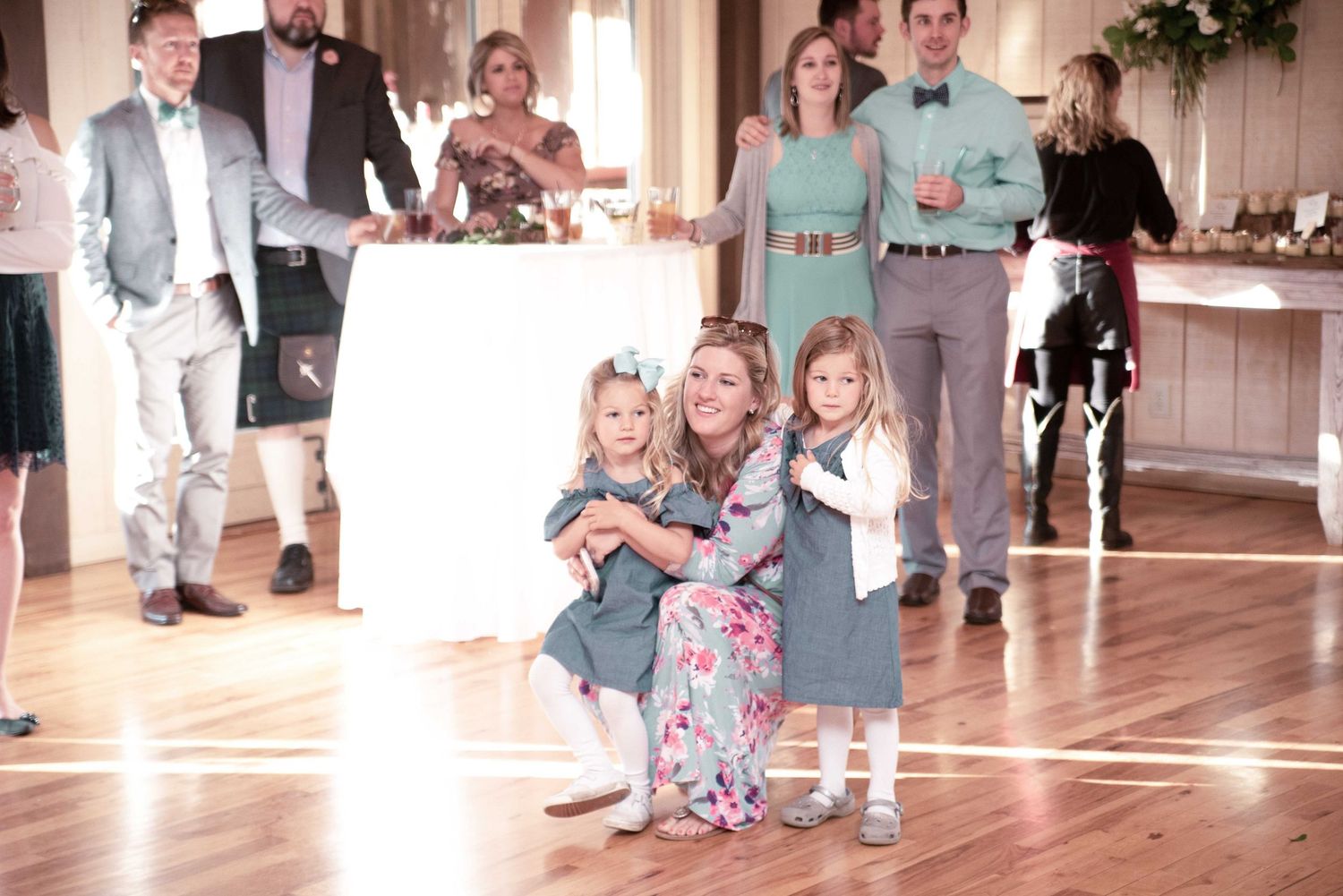 Woman in floral dress hugging two young girls in matching outfits on a wedding reception dance floor.