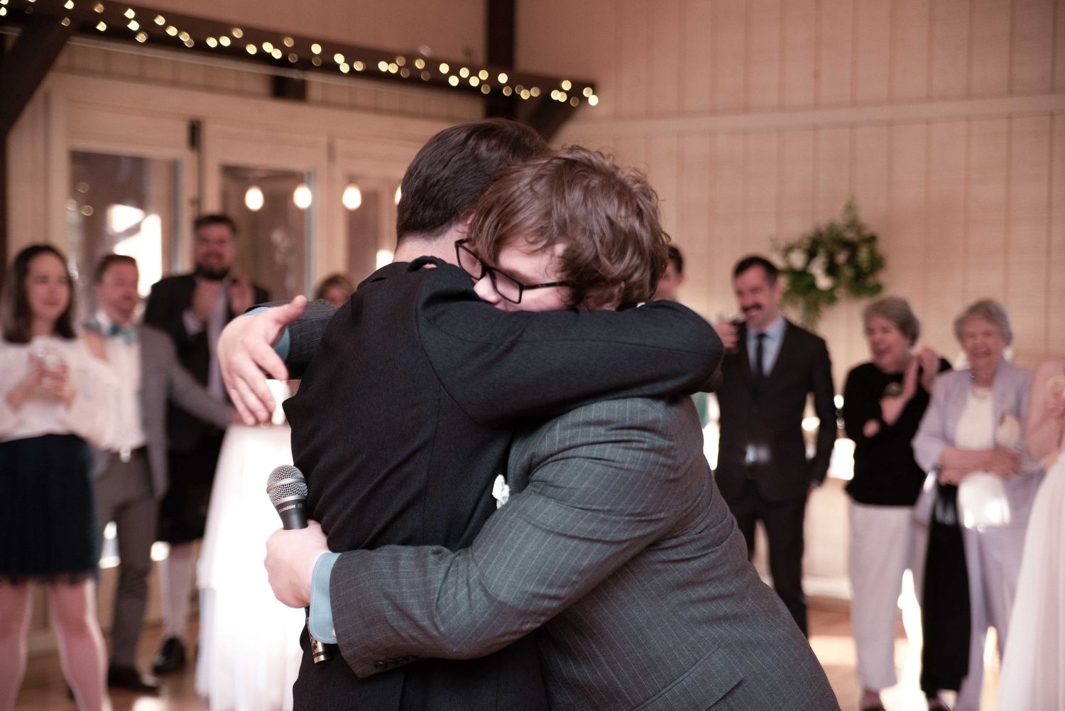 Two men share an emotional hug on a wedding dance floor surrounded by guests at a reception.