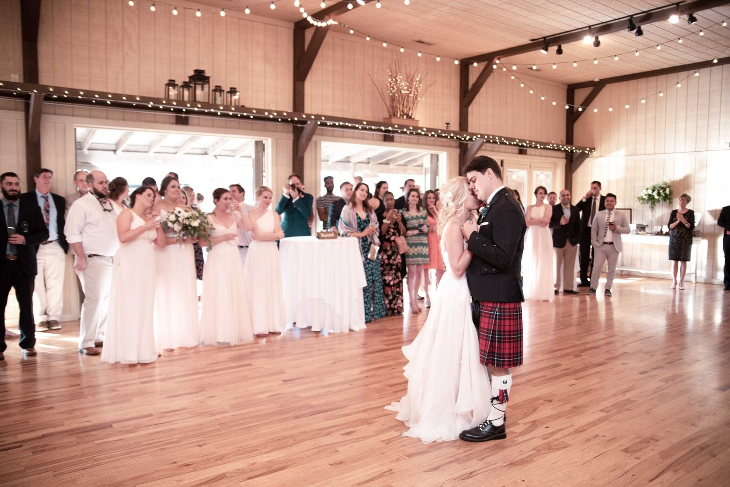 Bride and groom share first dance in rustic barn wedding venue with bridesmaids in blush gowns and guests watching.