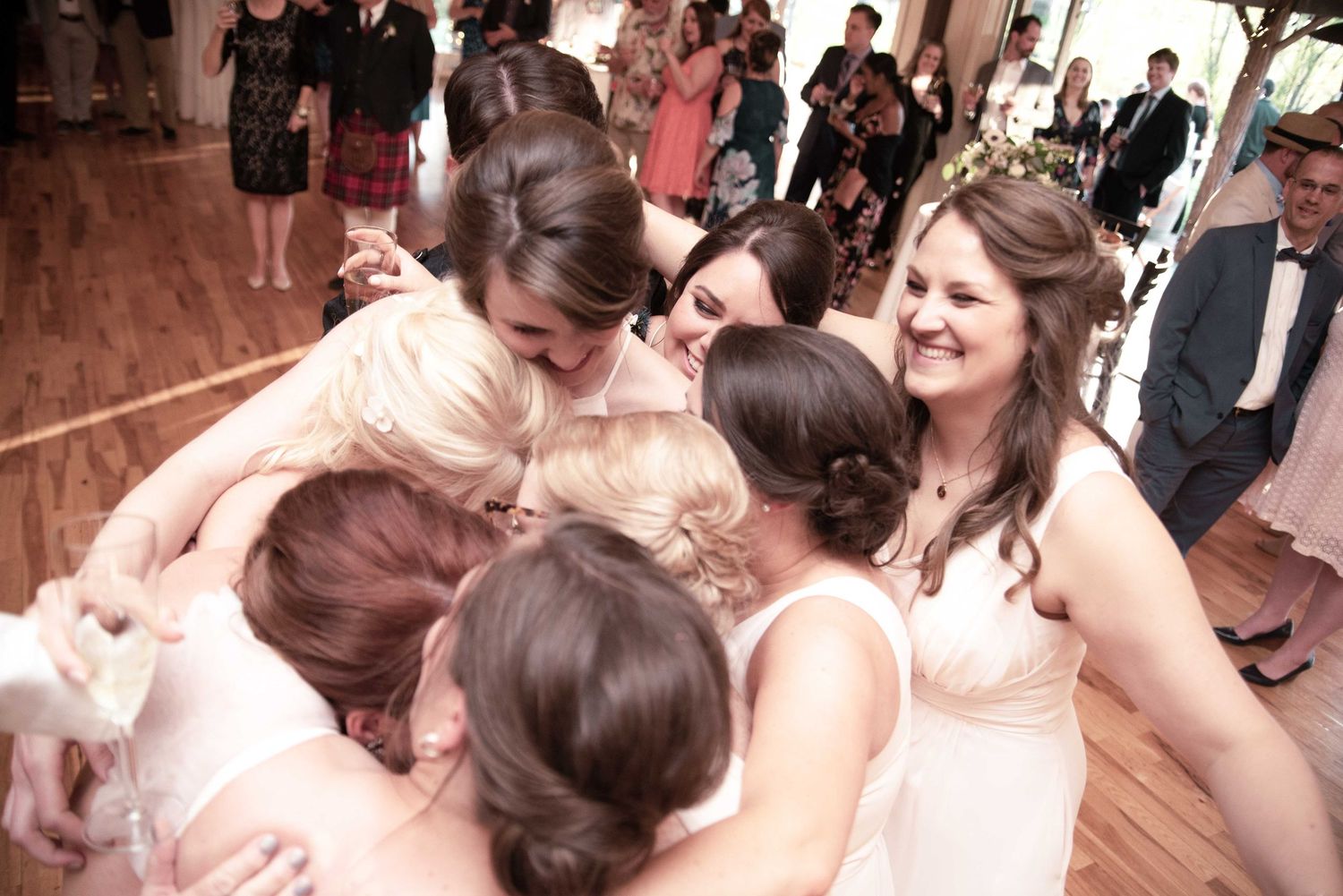 Bridesmaids in white dresses share a joyful group hug on the dance floor at a wedding reception.