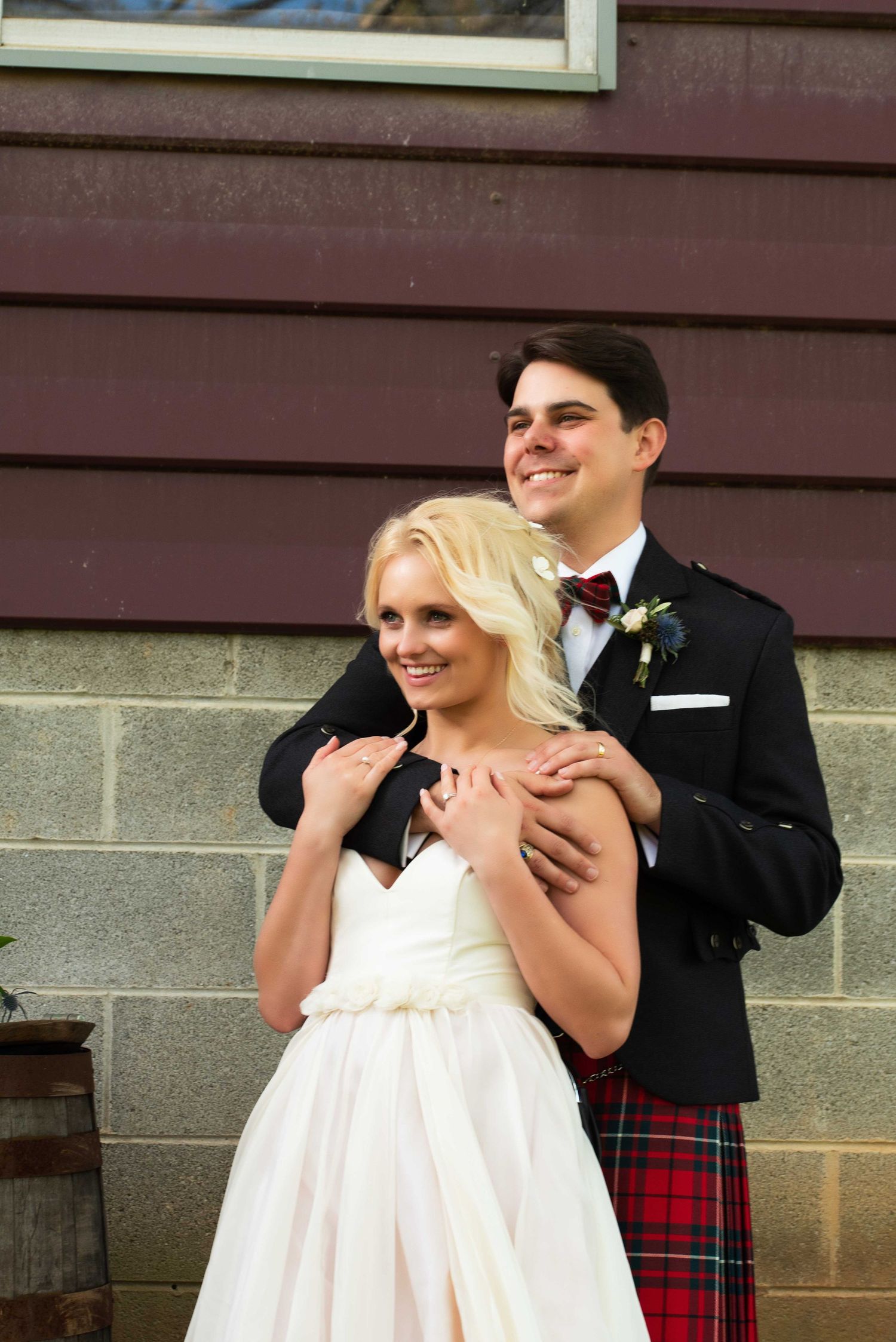 A smiling groom in a red tartan kilt embraces his blonde bride in a white wedding dress outdoors.