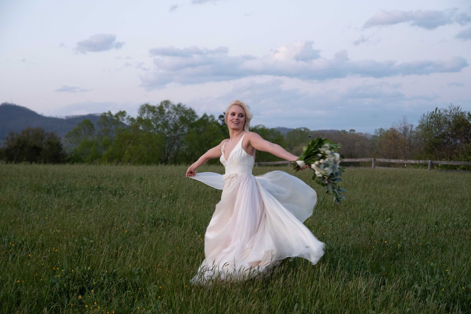 Bride in flowing white gown twirling in green field with mountain backdrop at dusk, holding bouquet.