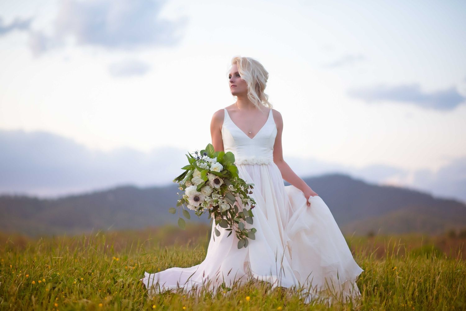 Bride in elegant white V-neck gown holds lush green and white bouquet in a scenic mountain meadow at sunset.
