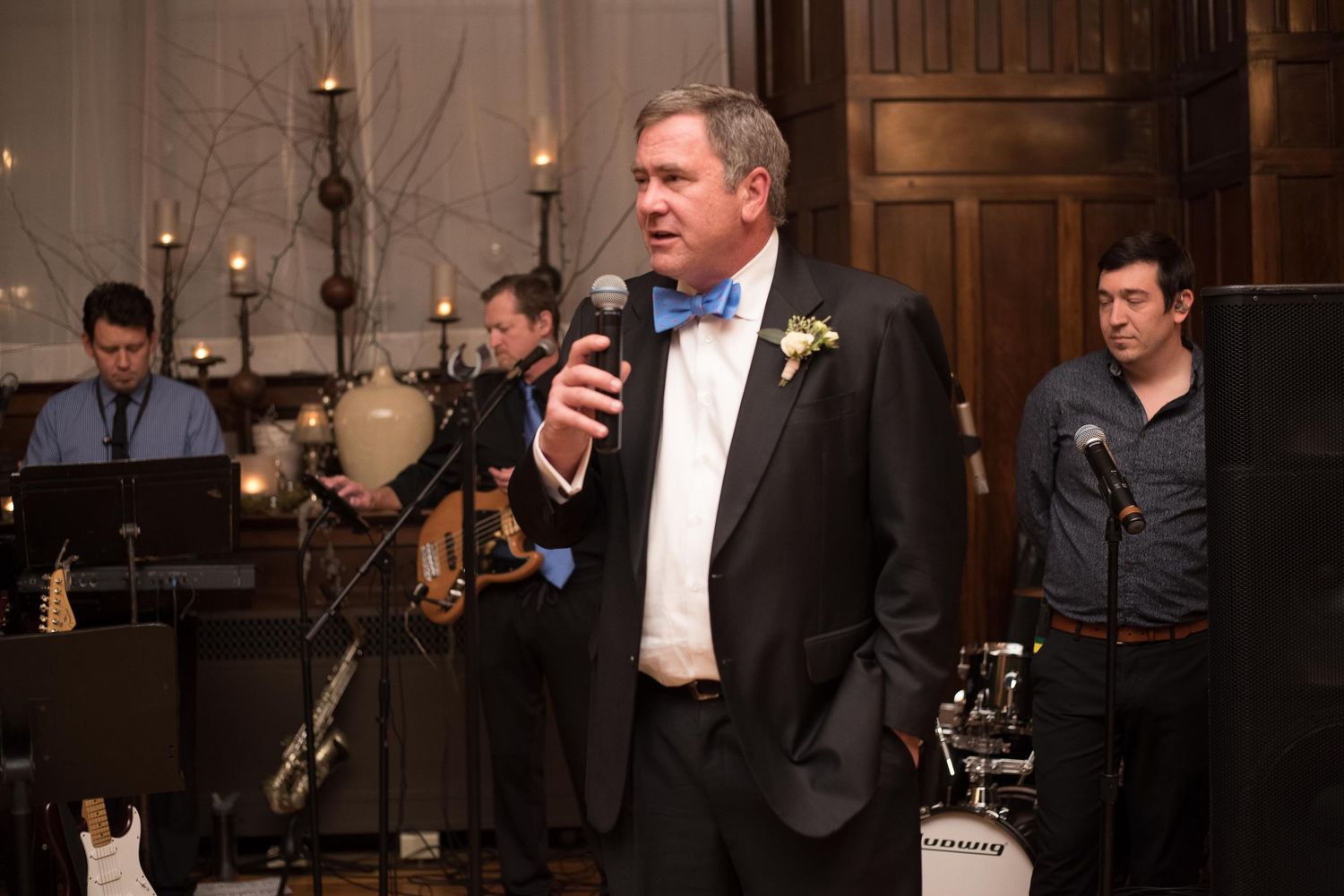 Man in tuxedo with blue bow tie speaks into microphone at elegant wedding reception with live band.