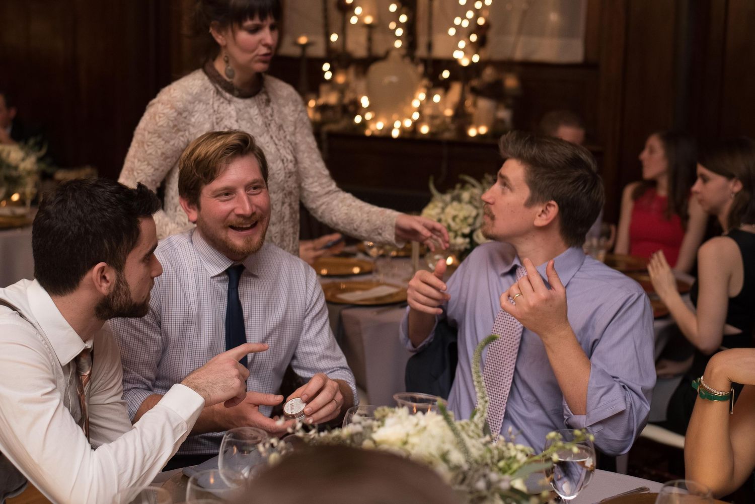 Three guests laughing and chatting at an elegant wedding reception dinner with warm string lights in background.