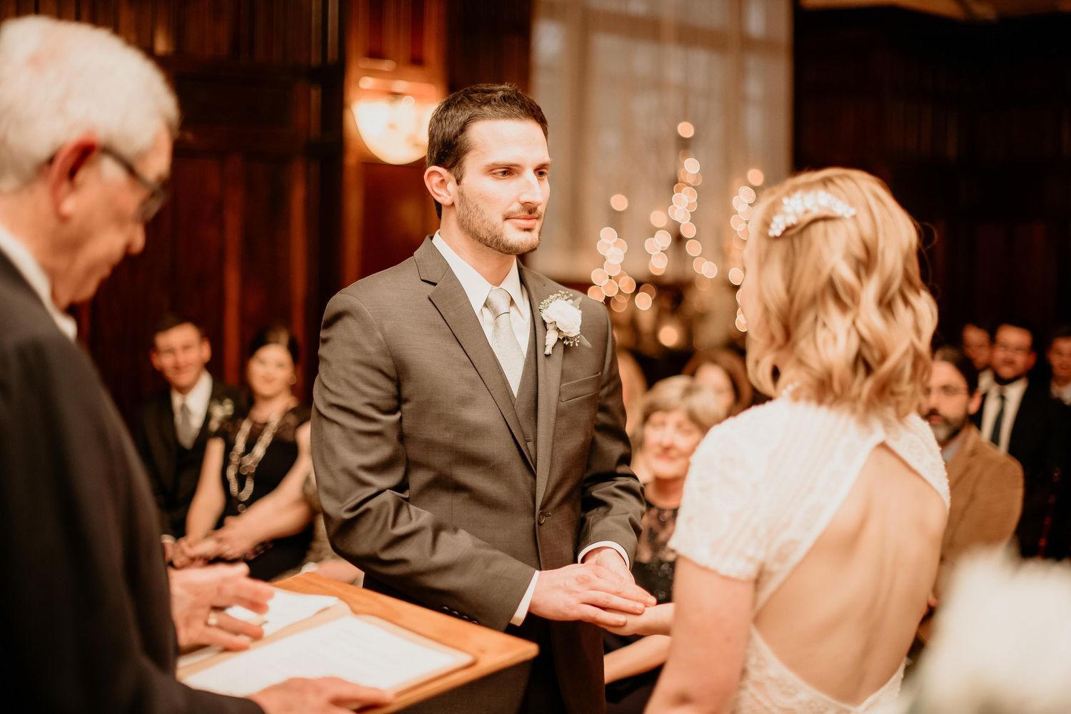 Groom in gray suit exchanges vows with bride at elegant indoor wedding ceremony with warm lighting.