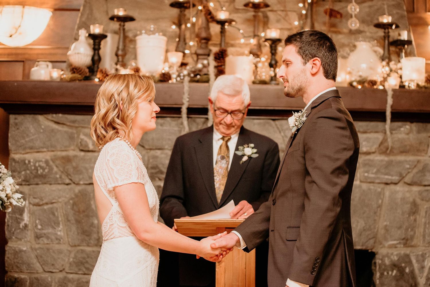 Couple exchanging vows at rustic stone fireplace wedding ceremony with officiant and candle backdrop.