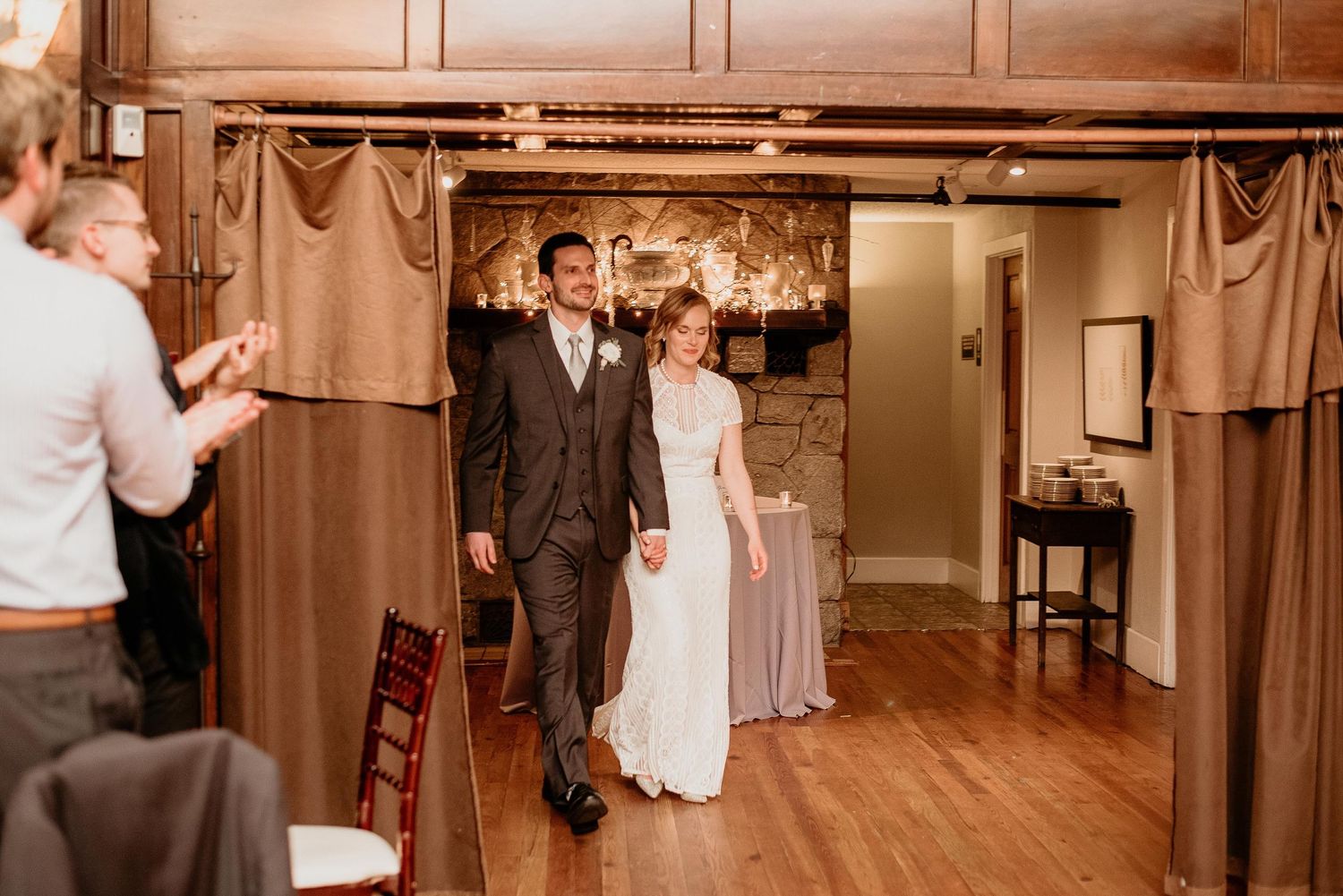 Bride and groom make grand entrance at rustic wedding reception venue with warm wood accents and stone fireplace.
