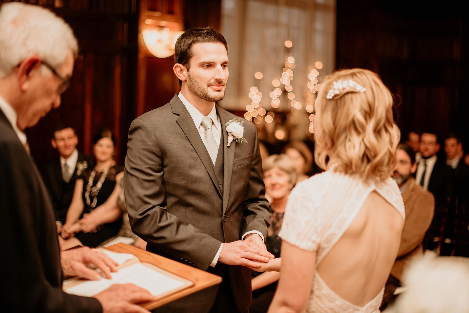 Groom in gray suit smiling at bride during indoor wedding ceremony with guests and warm bokeh lights in background.