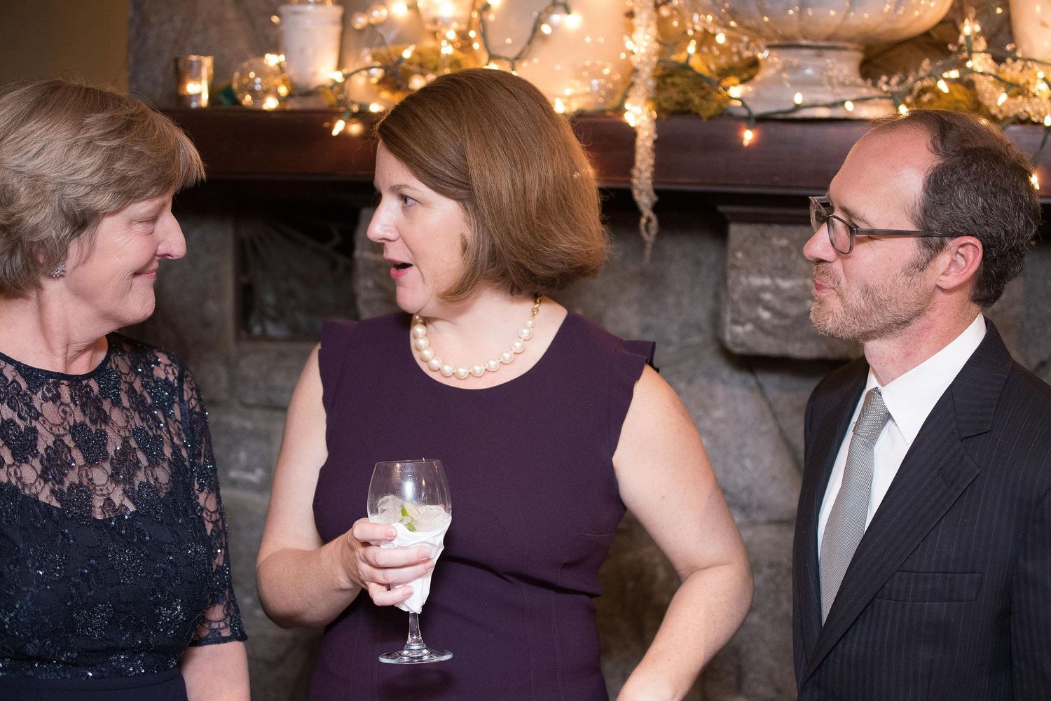 Three adults socializing at an elegant holiday party near a decorated fireplace, woman holding a wine glass.