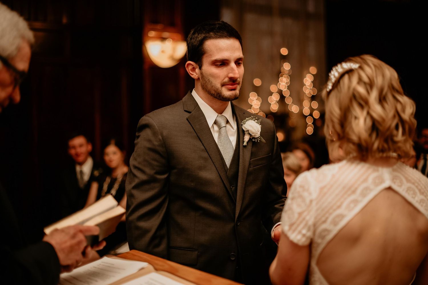 Groom in gray suit gazing at bride during indoor wedding ceremony with warm bokeh lights in background.