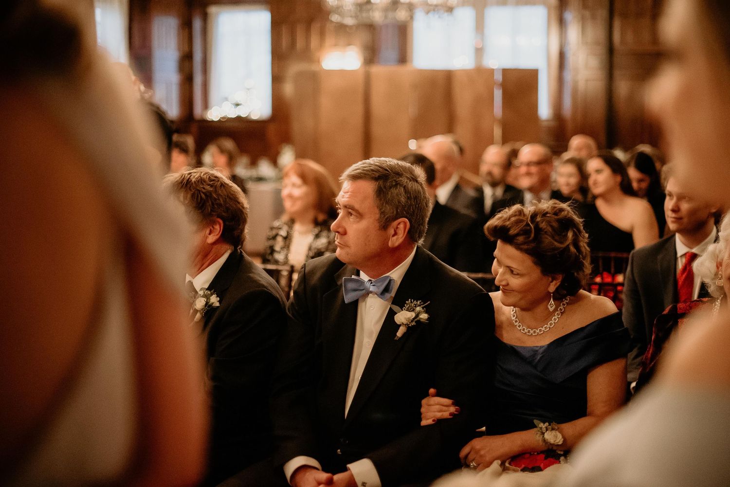 Guests seated at a wedding ceremony in an elegant wood-paneled hall, man in tuxedo with bow tie smiling warmly.