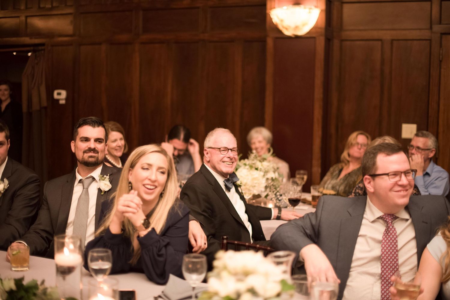 Guests laughing and enjoying themselves at an elegant formal wedding reception dinner in a wood-paneled ballroom.