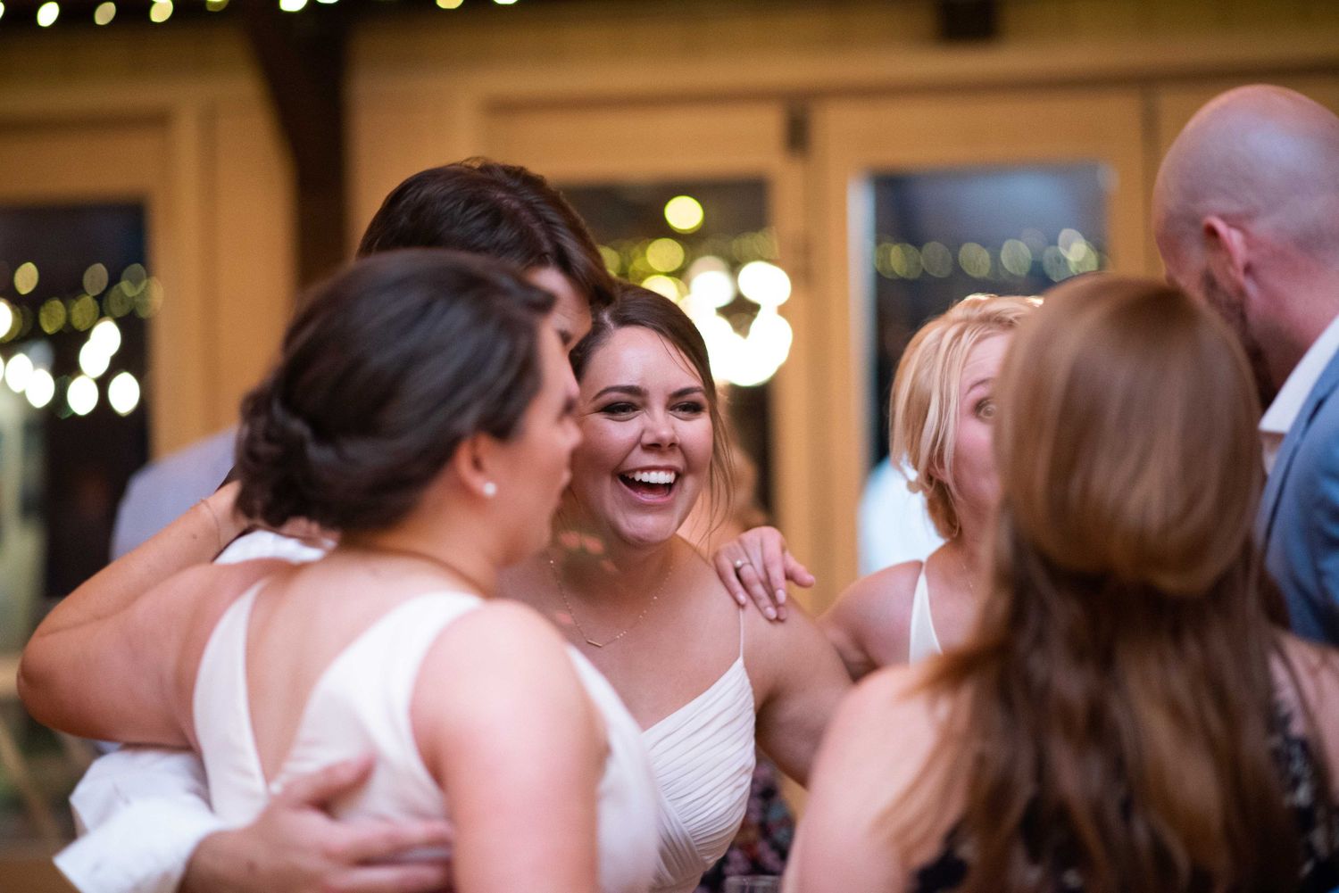 Bride laughing joyfully with bridesmaids during wedding reception dance floor celebration indoors with warm string lights.