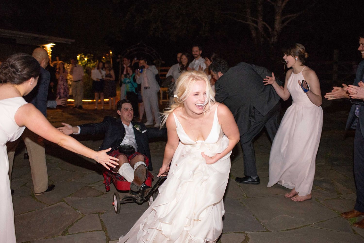 Bride in white gown dancing energetically at outdoor wedding reception with guests celebrating at night.