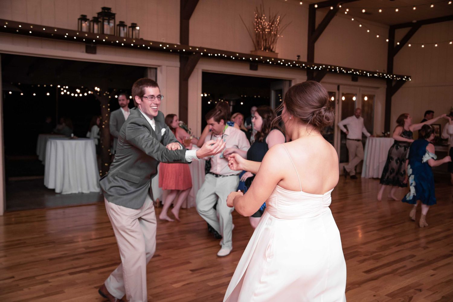 Guests dancing joyfully at a rustic barn wedding reception with string lights and hardwood floors.