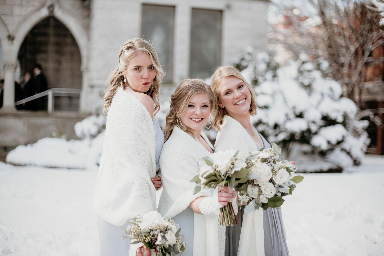 Three women in white wraps holding bouquets pose in snow outside a church on a winter wedding day.