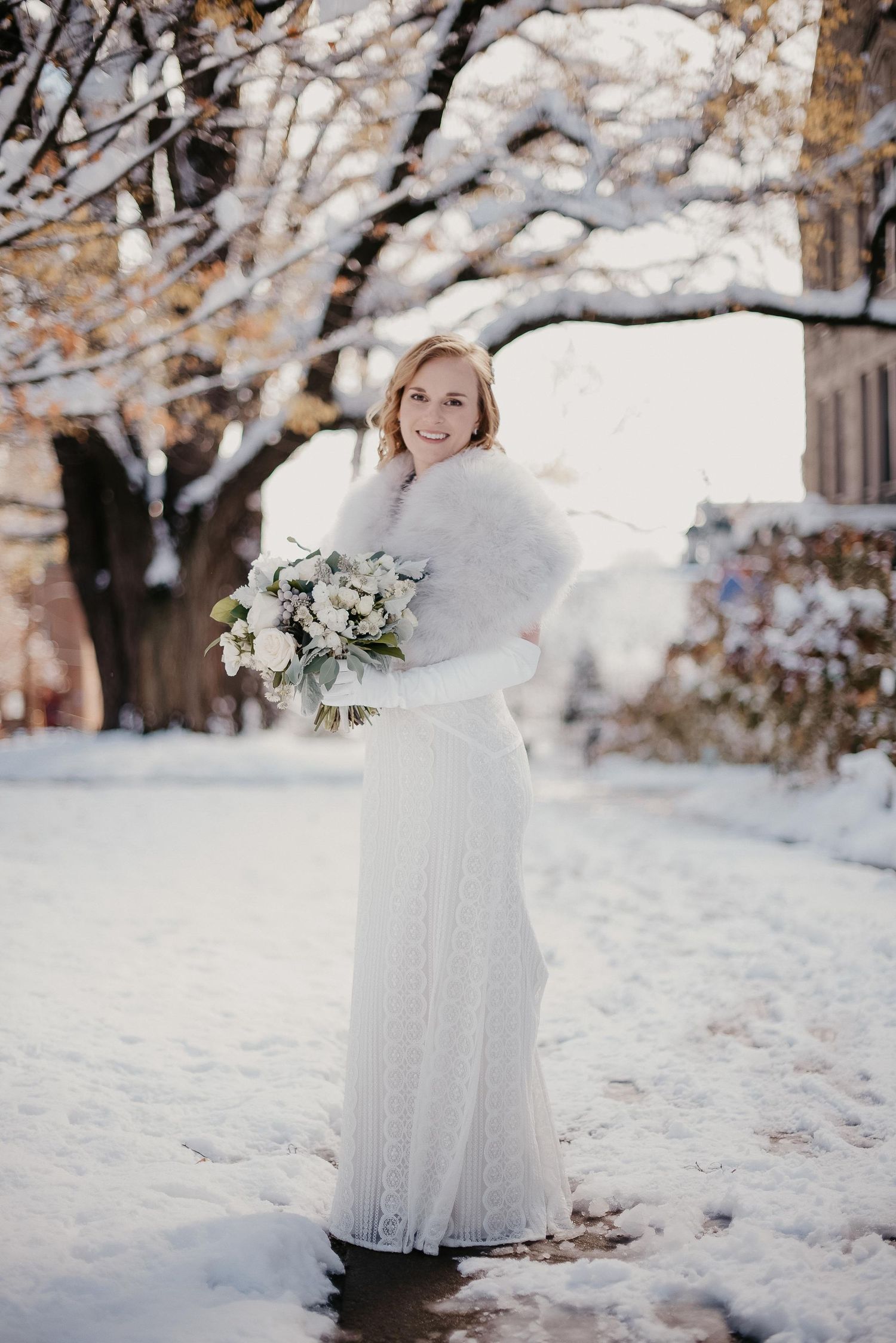 Bride in white lace gown and fur stole holding bouquet, smiling in snowy winter outdoor setting.