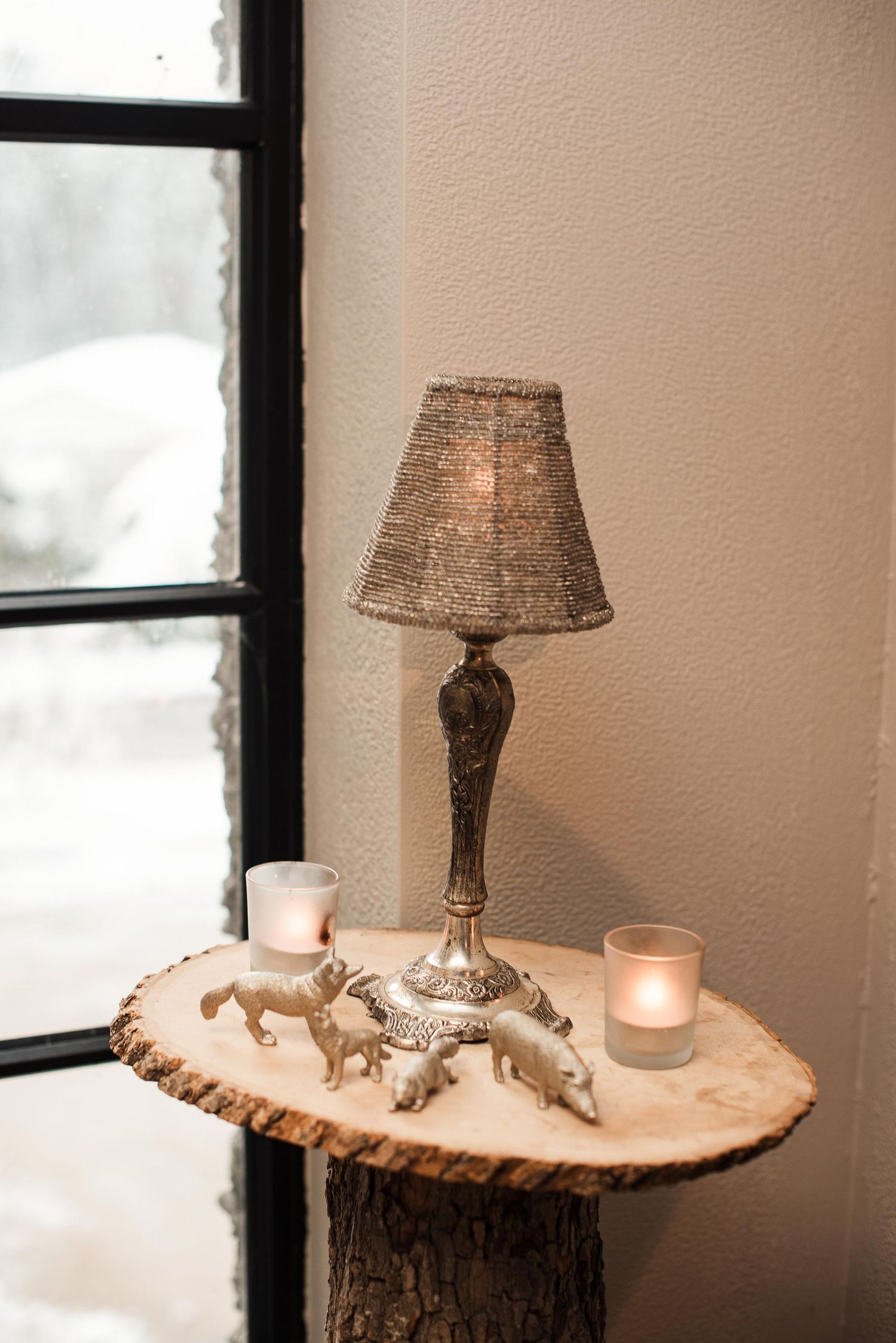 Small ornate silver lamp with burlap shade on wood slice table beside lit candles and silver animal figurines near window.