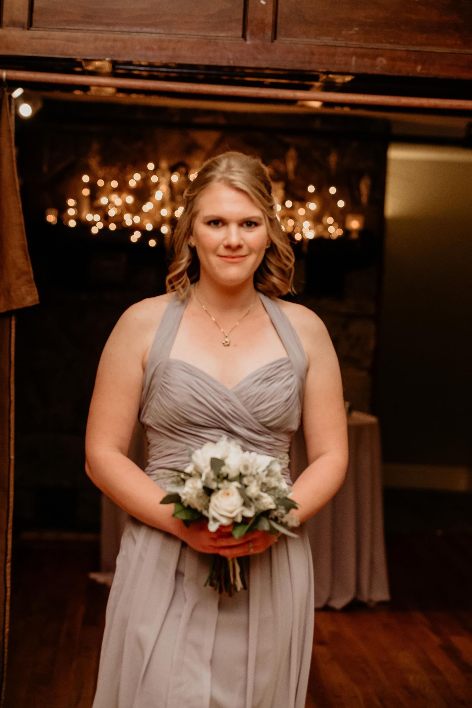 Bridesmaid in gray halter gown holding white bouquet stands before warm string lights at elegant wedding venue.