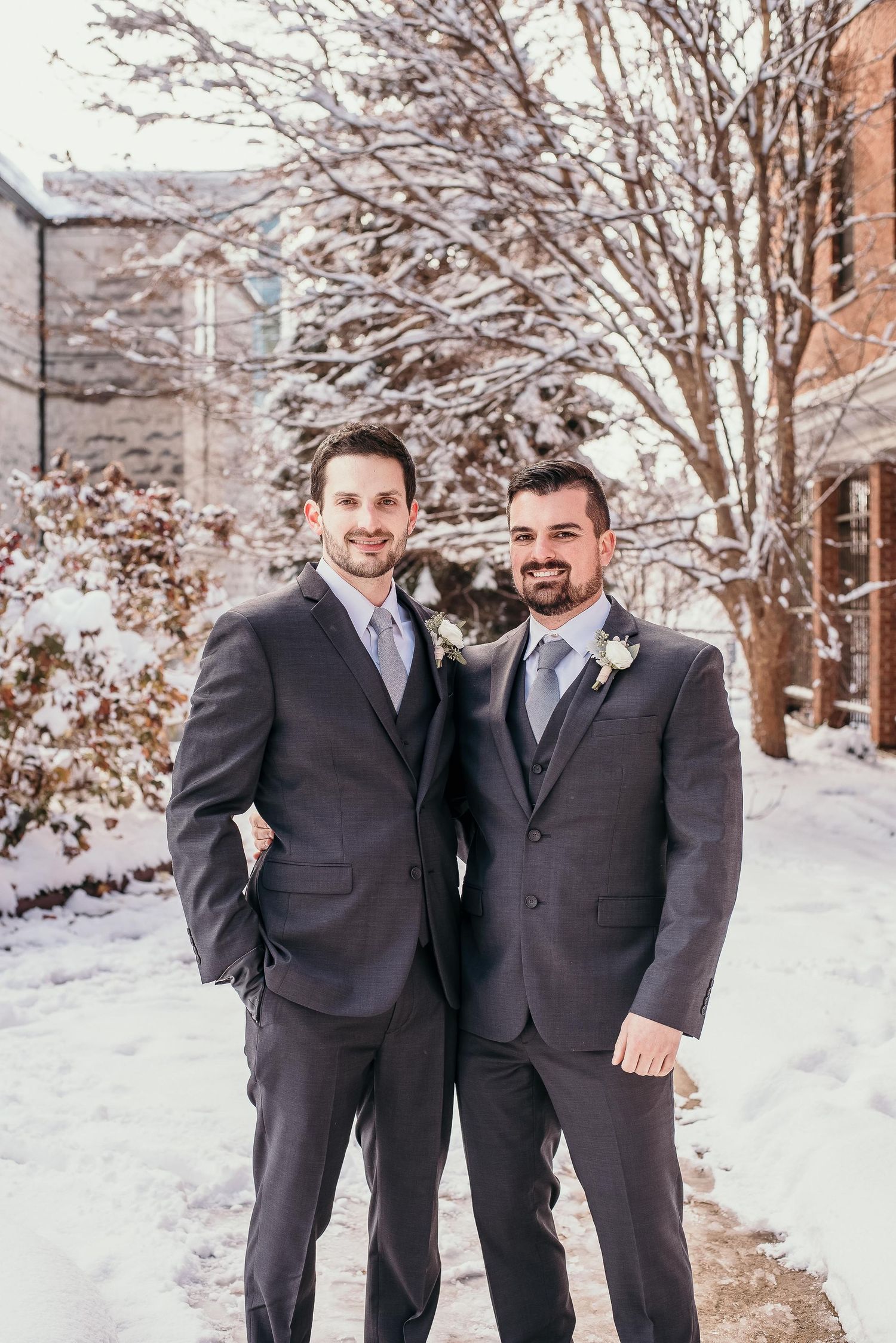 Two grooms in matching gray suits pose outdoors in a snowy winter wedding setting with snow-covered trees.