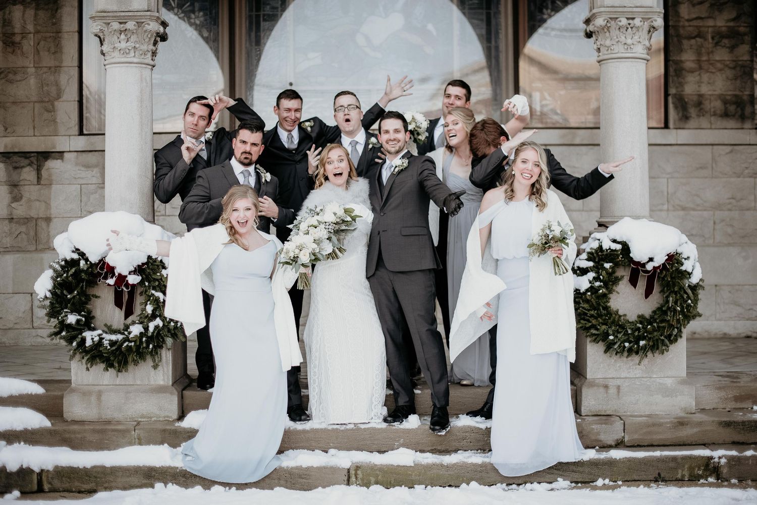 Winter wedding party posing on snow-covered steps with holiday wreaths, bride and bridesmaids in white, groomsmen in gray suits.