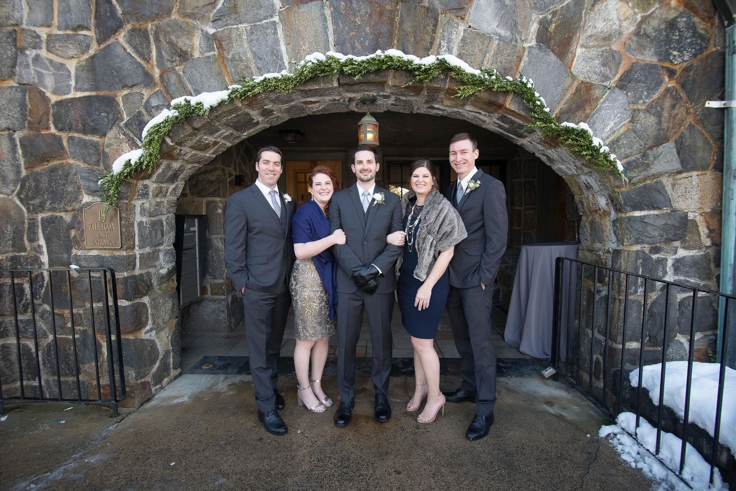 Five elegantly dressed people posing under a stone archway decorated with greenery at a winter wedding venue.